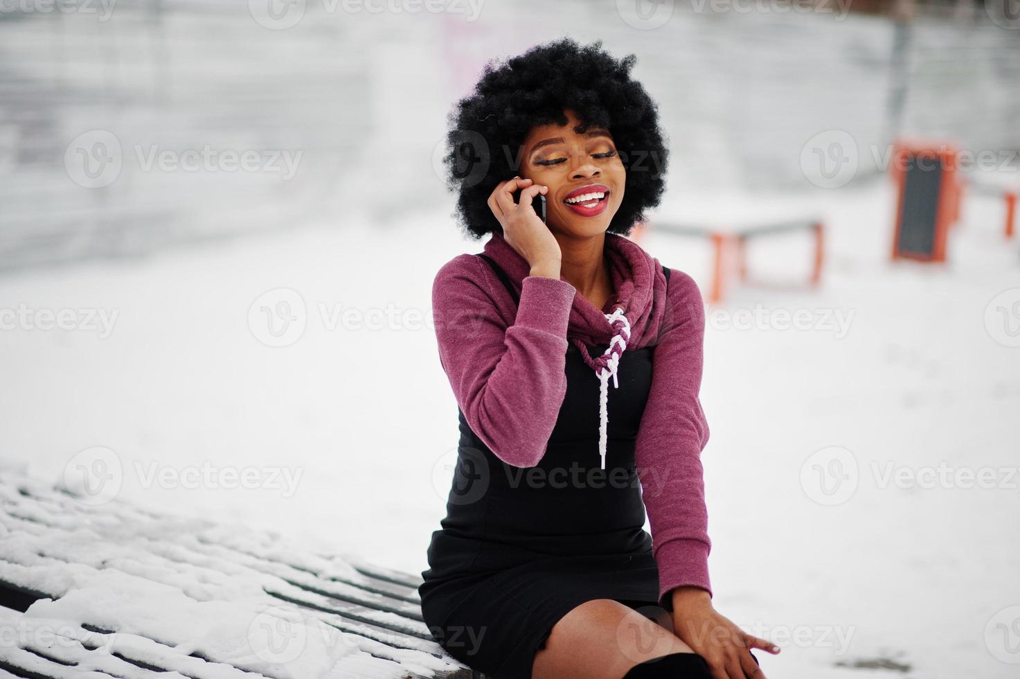 Curly hair african american woman posed at winter day, sitting on bench and speaking on mobile phone. photo