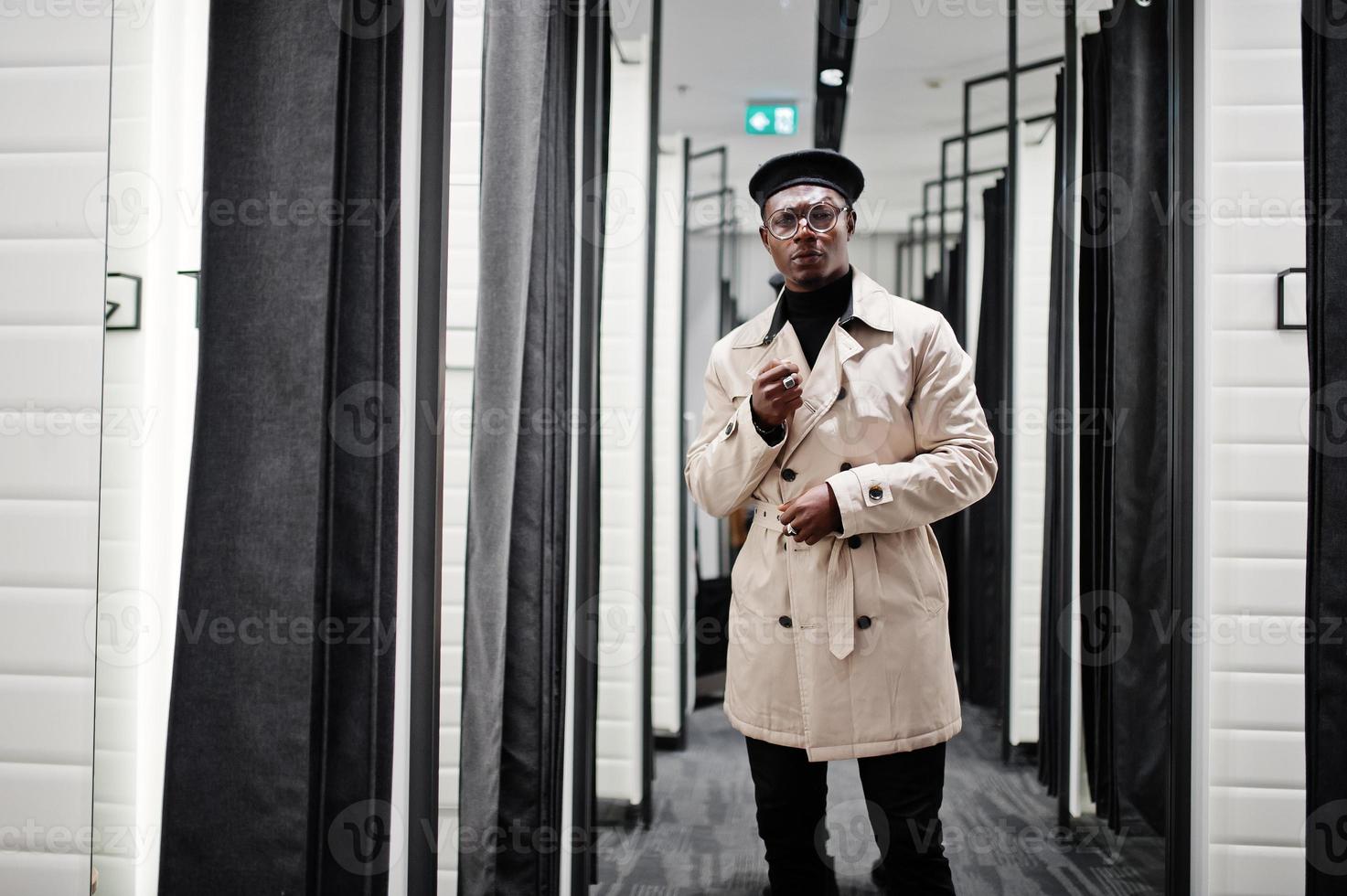 Elegante hombre afroamericano casual con boina negra y impermeable de trinchera blanca en la tienda de ropa de la sala de montaje. foto