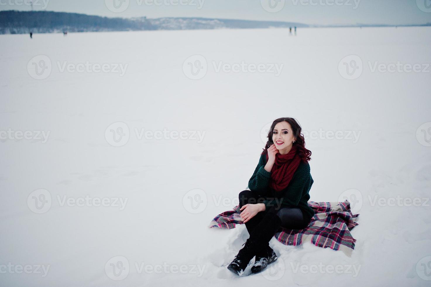 chica morena con suéter verde y bufanda roja sentada en un lago congelado al aire libre a cuadros en el día de invierno por la noche. foto