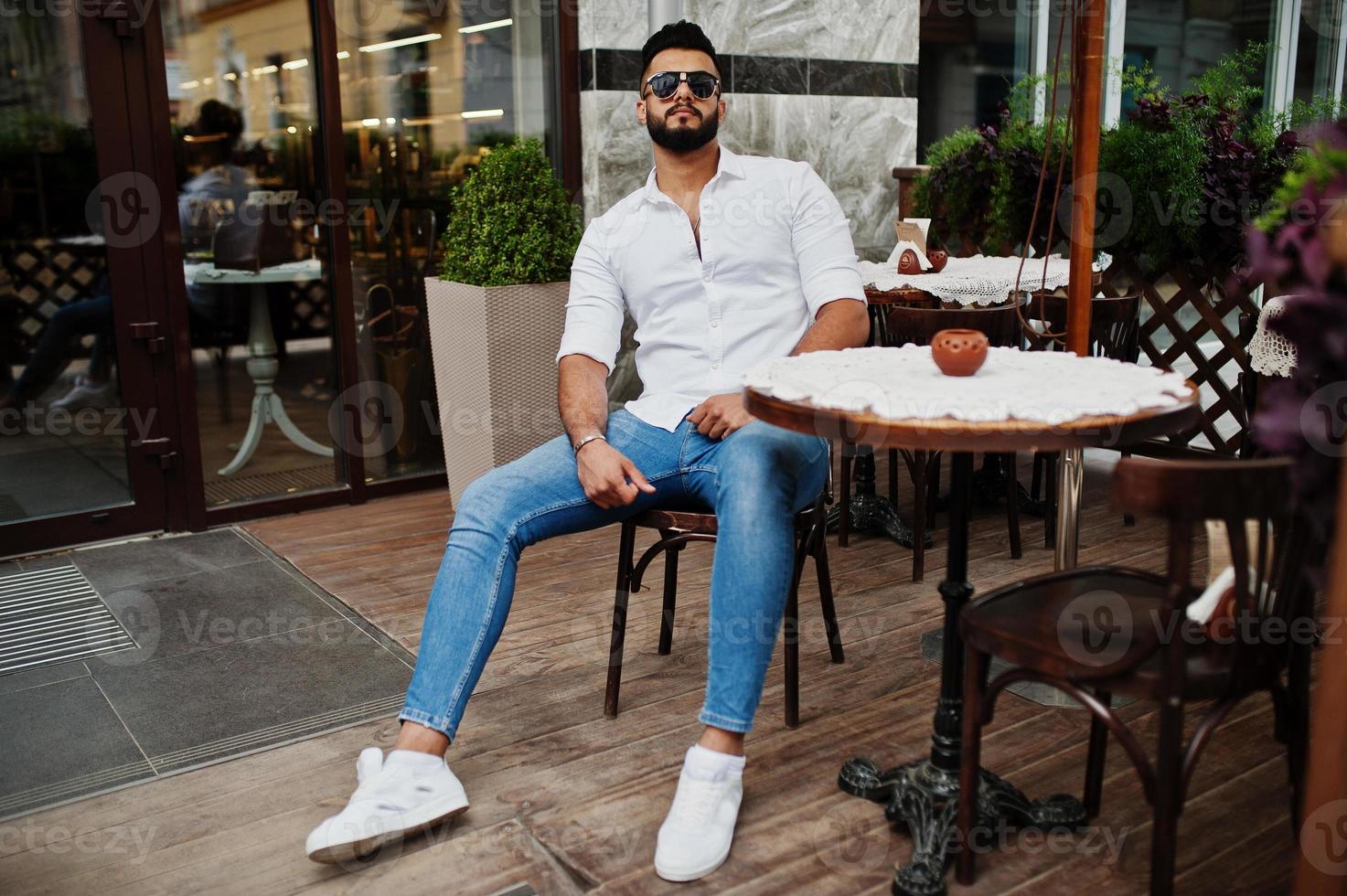 Stylish tall arabian man model in white shirt, jeans and sunglasses posed at street of city. Beard attractive arab guy sitting on outdoor cafe. photo