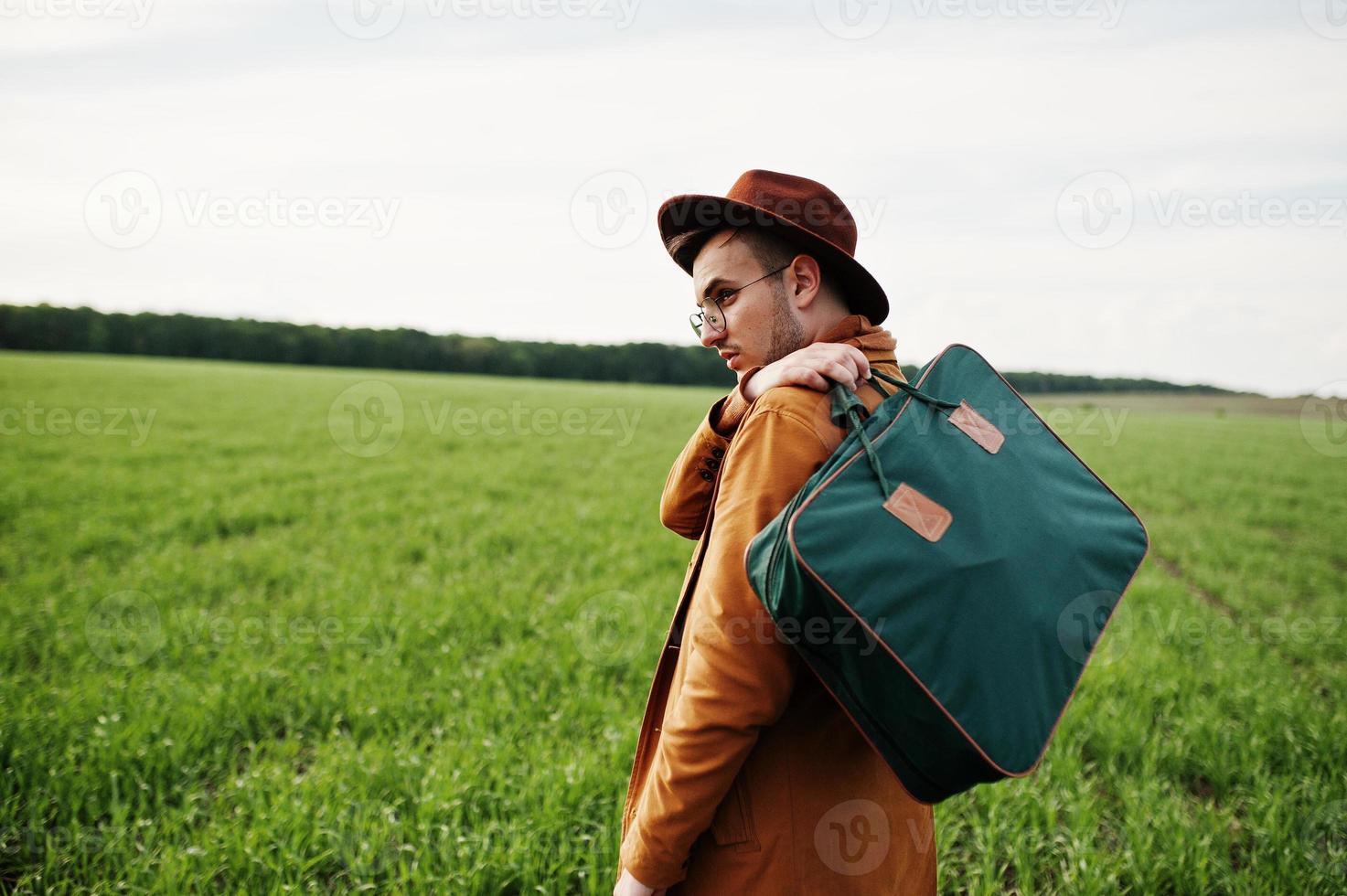 Stylish man in glasses, brown jacket and hat with bag posed on green field. photo