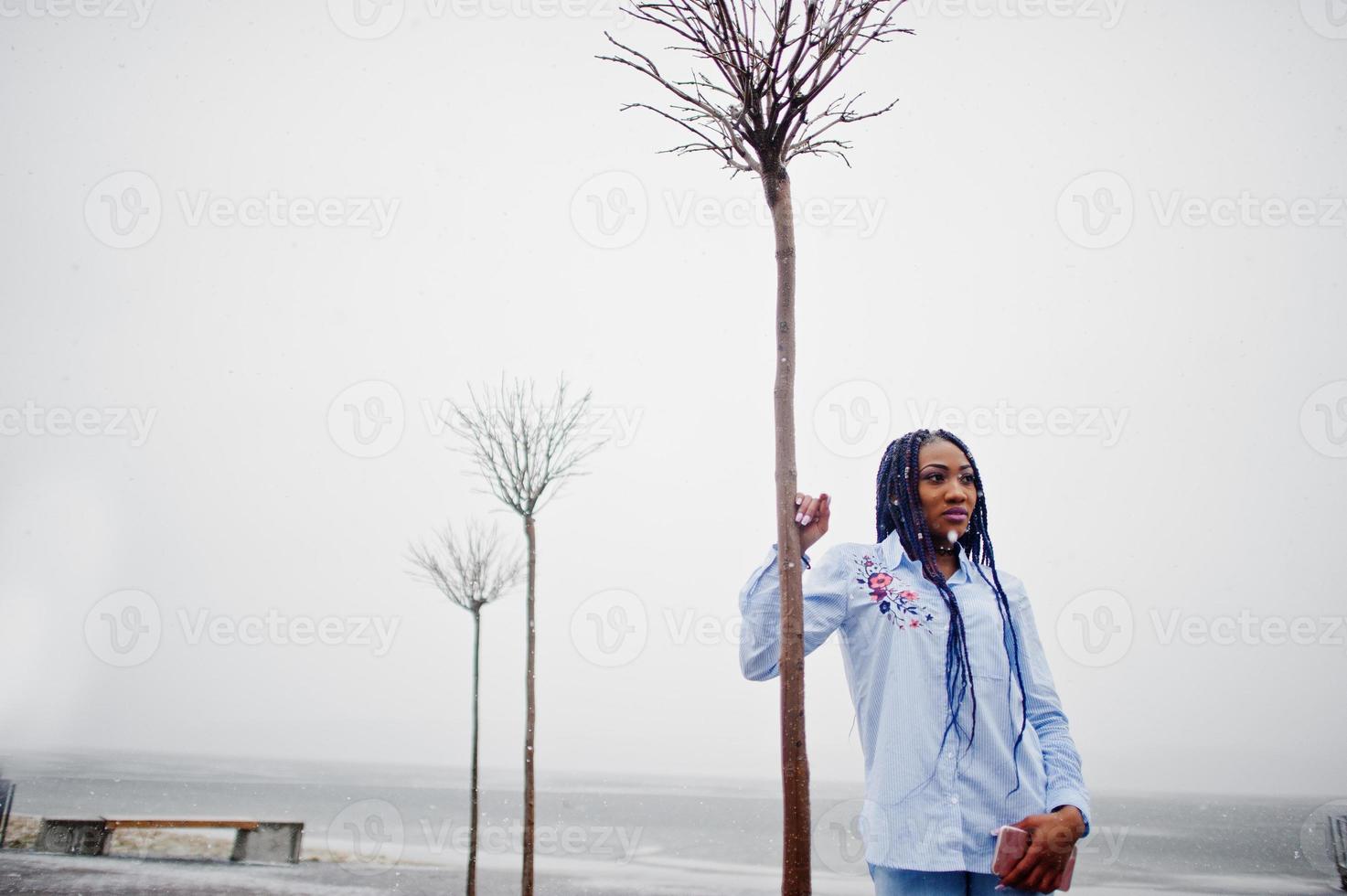 Stylish african american girl with dreads holding mobile phone at hand, outdoor on pier against frozen lake at snowy weather. photo