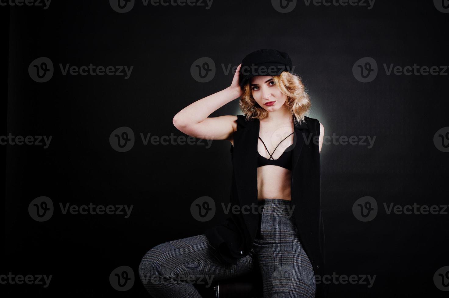 Studio portrait of blonde girl in black wear, bra and cap against dark background. photo