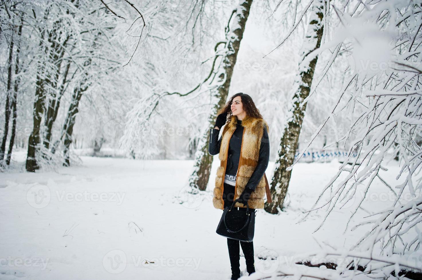 Elegance curly girl in fur coat and handbag at snowy forest park at winter. photo