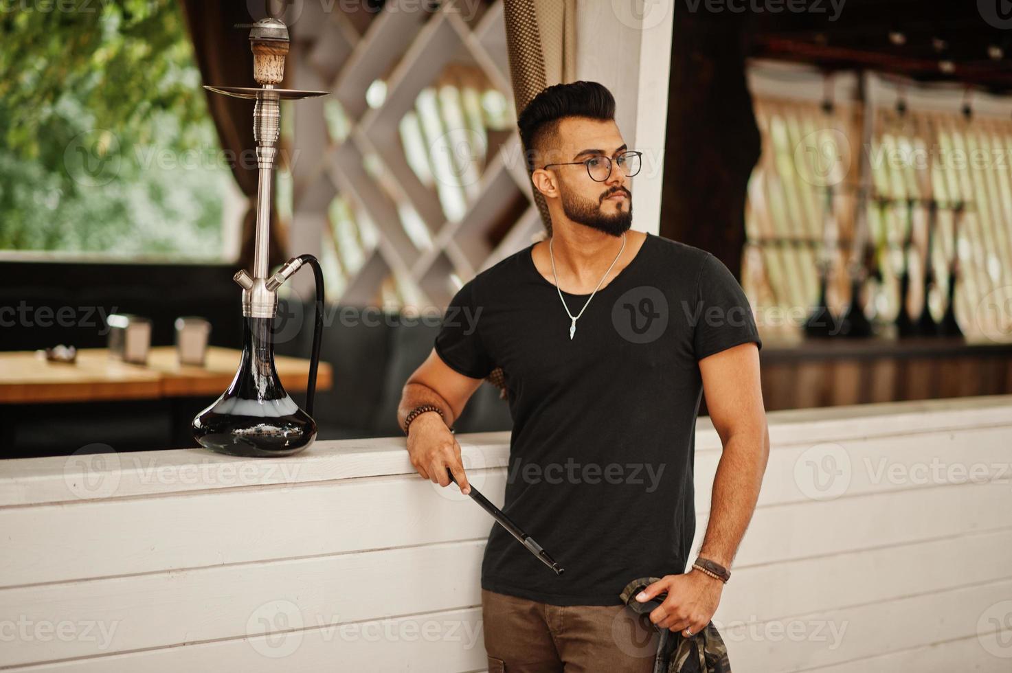 Stylish beard arabian man in glasses and black t-shirt smoking hookah outdoor. Arab model having rest. photo