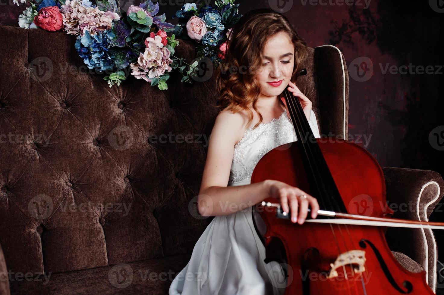 Pretty young gilrl musician in white dress with double bass sitting on brown vintage sofa. photo