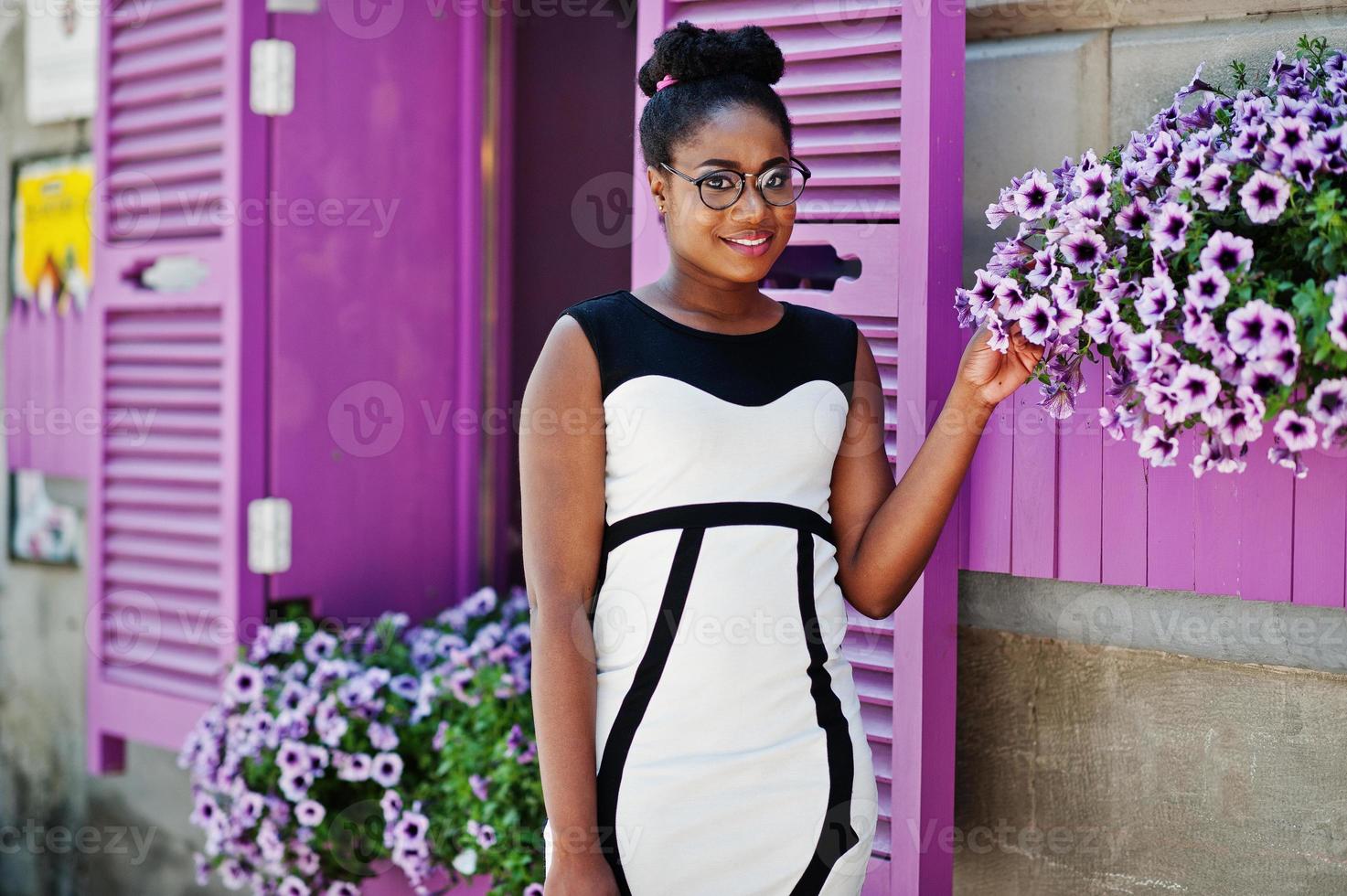 chica afroamericana con gafas, vestido blanco y negro y bolso posado contra ventanas moradas. foto
