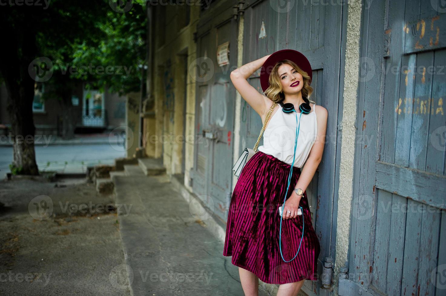 Fashionable and beautiful blonde model girl in stylish red velvet velour skirt, white blouse and hat, posed with phone and earphones. photo