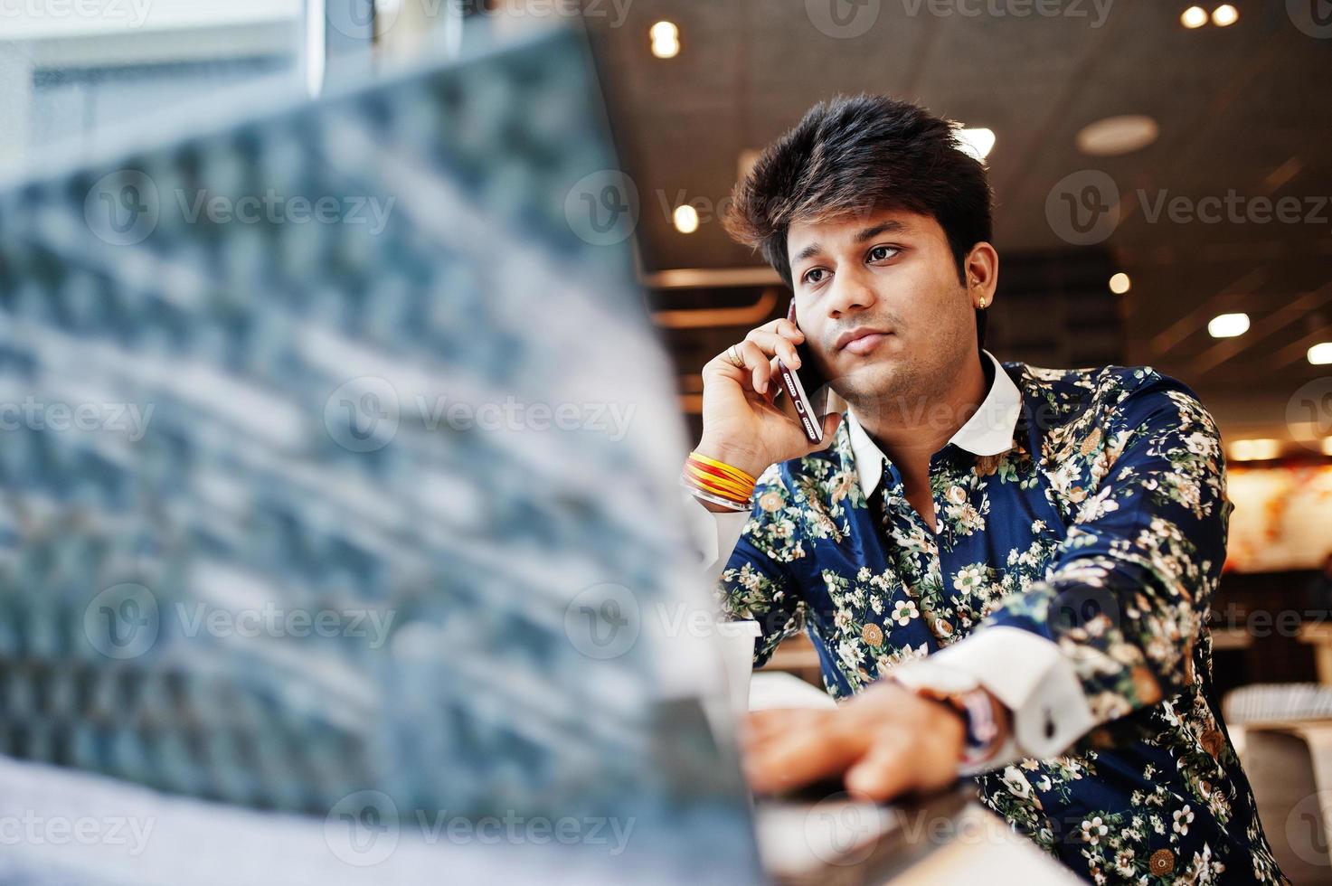 Young male indian freelancer sitting open laptop computer in fast food cafe, handsome asian man speaking phone during work on net-book in comfortable coffee shop. photo