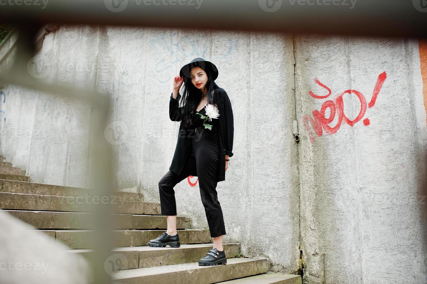 chica sensual todo en negro, labios rojos y sombrero. mujer gótica dramática sostiene una flor de crisantemo blanco contra la pared de graffiti. foto
