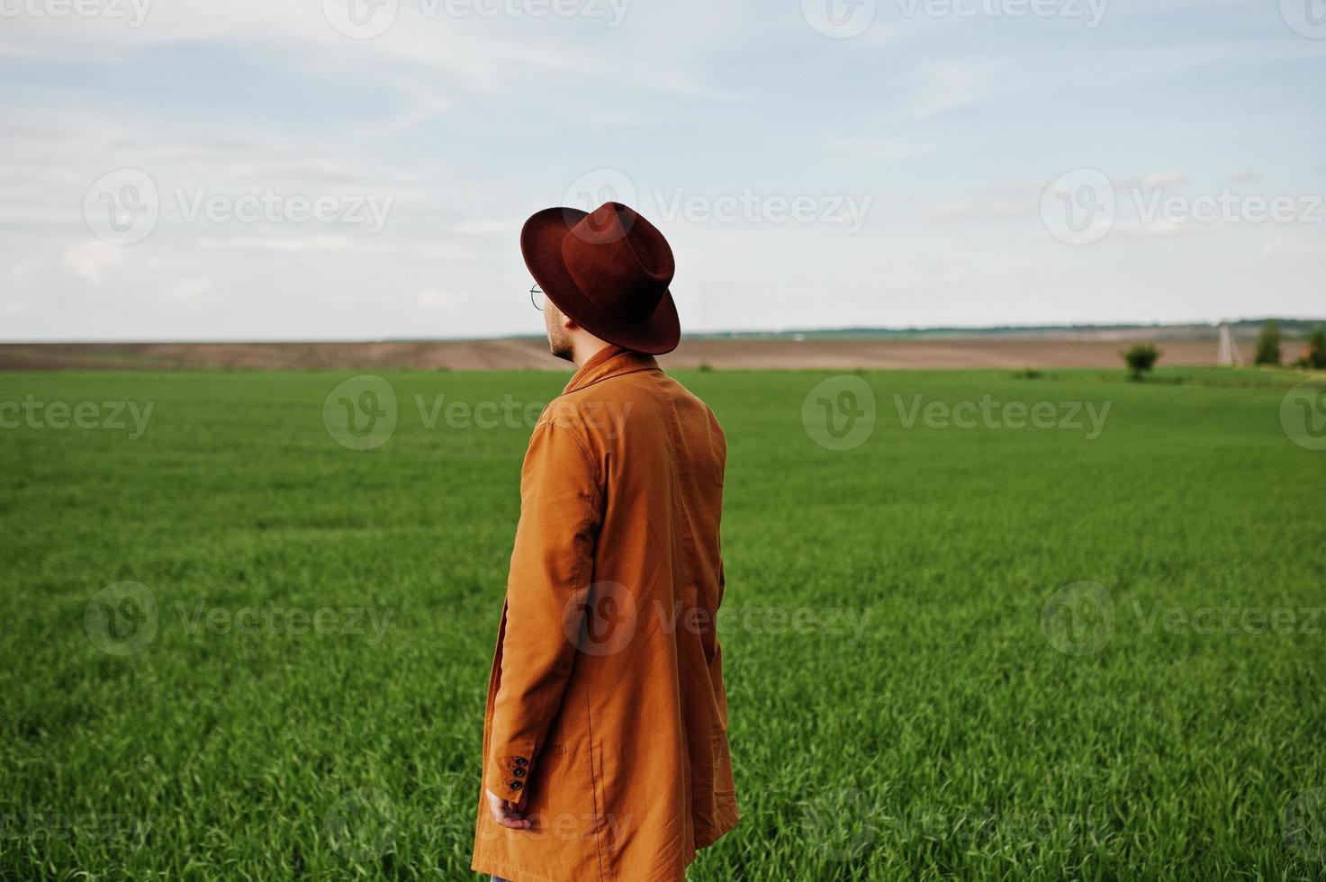 hombre elegante con gafas, chaqueta marrón y sombrero posado en campo verde. foto