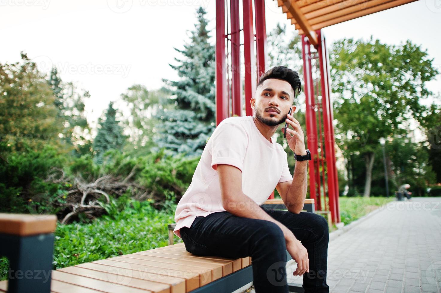 Stylish indian beard man at pink t-shirt. India model sitting on bench of city and use his mobile phone. photo