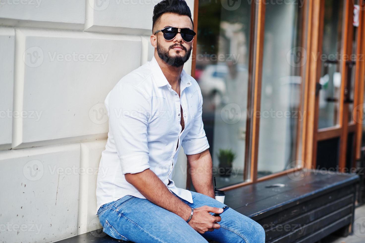 Stylish tall arabian man model in white shirt, jeans and sunglasses posed at street of city and hold mobile phone at hand. Beard arab guy with cup of coffee sitting and having rest. photo