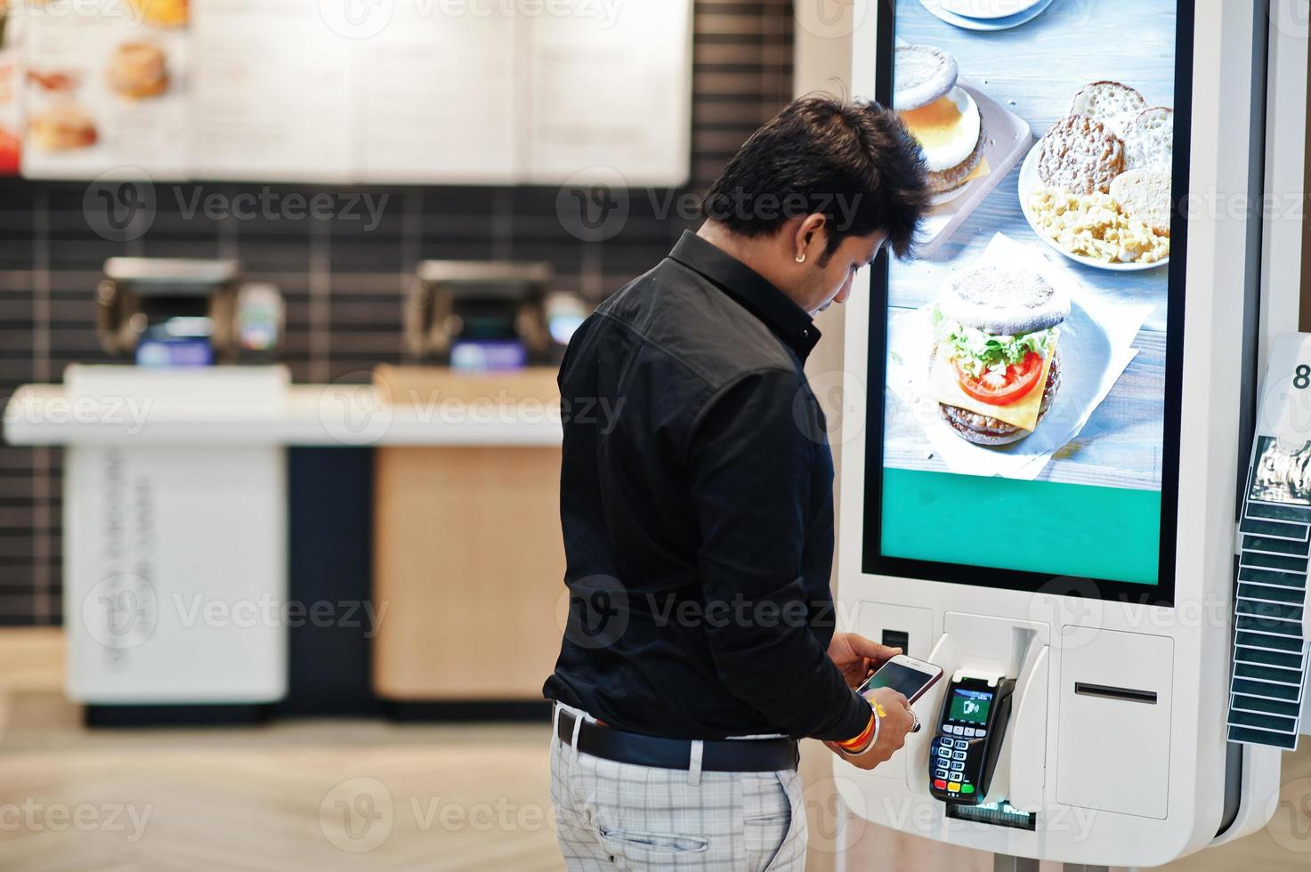 Indian man customer at store place orders and pay by contactless credit card on mobile phone through self pay floor kiosk for fast food, payment terminal. Pay pass. photo