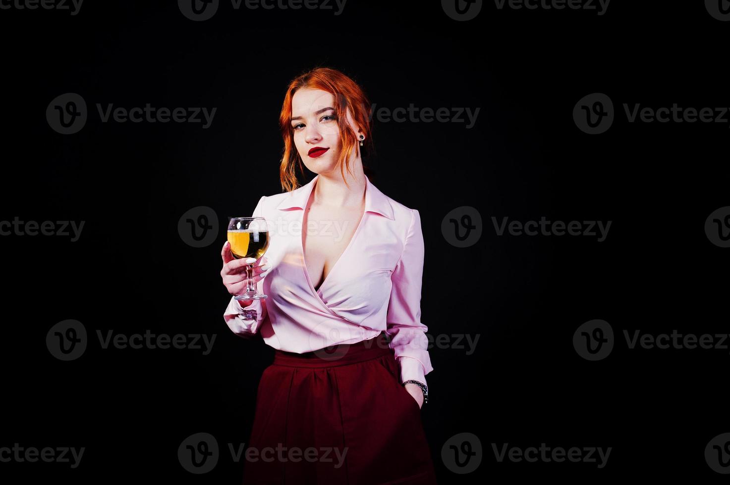 Gorgeous red haired girl in pink blouse and red skirt with glass of wine at hand isolated on black. photo