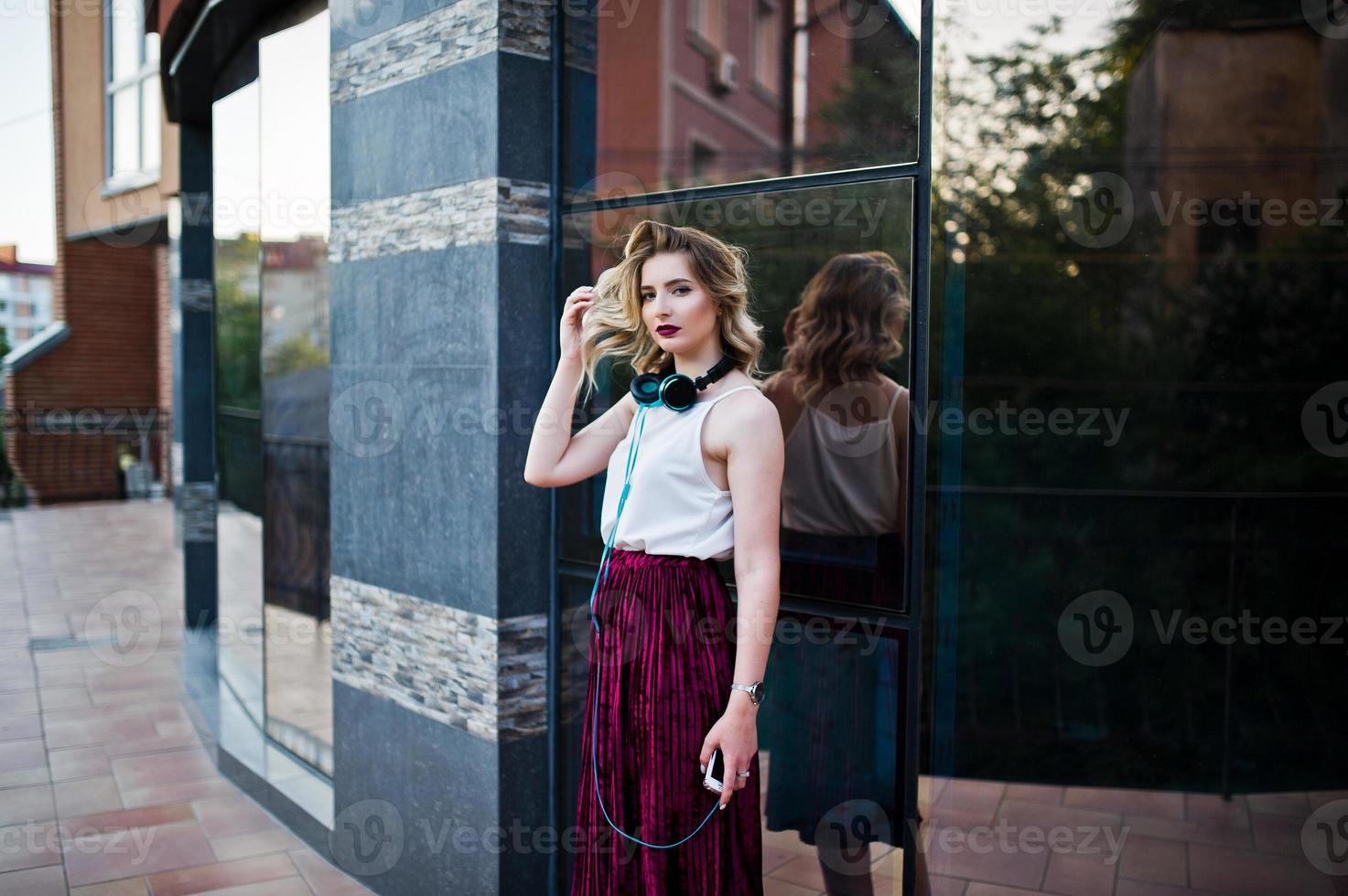 Fashionable and beautiful blonde model girl in stylish red velvet velour skirt, white blouse, posed with phone and earphones against windows of city building. photo