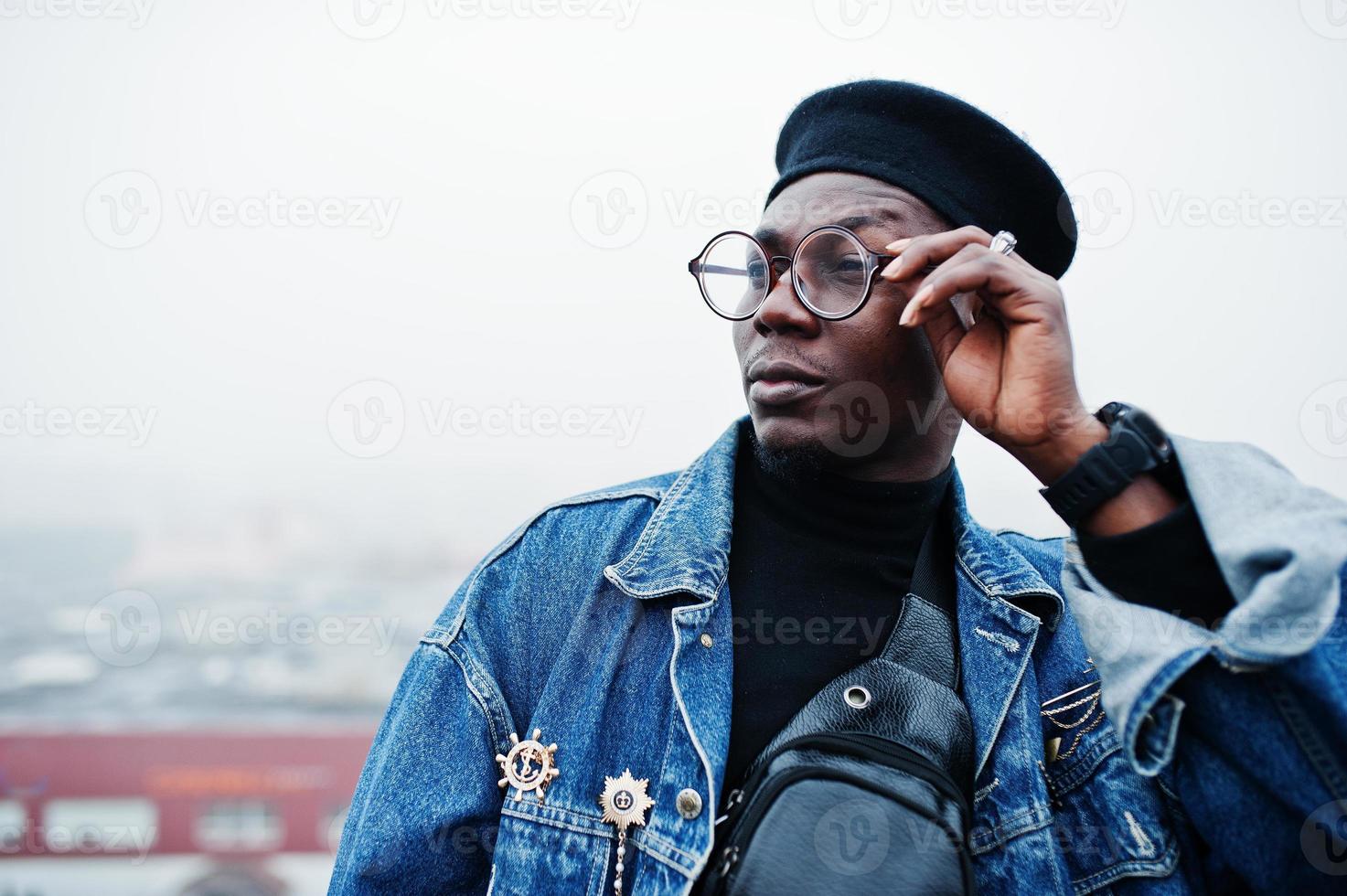 hombre afroamericano con chaqueta de jeans, boina y anteojos posados en un techo abandonado. foto