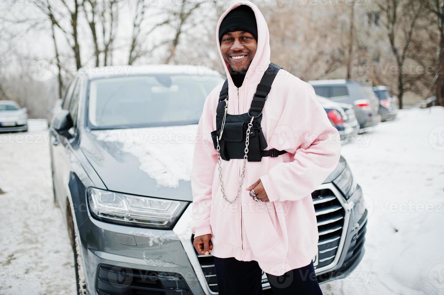 Stylish urban style african american man in pink hoodie posed against suv car at winter. Afro rapper guy. photo