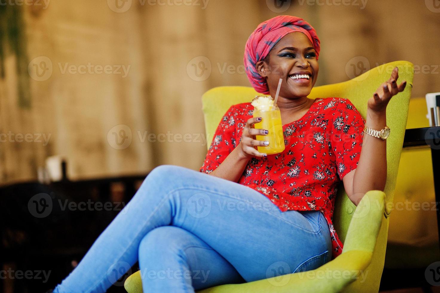 Stylish african woman in red shirt and hat posed indoor cafe and drinking pineapple lemonade. photo