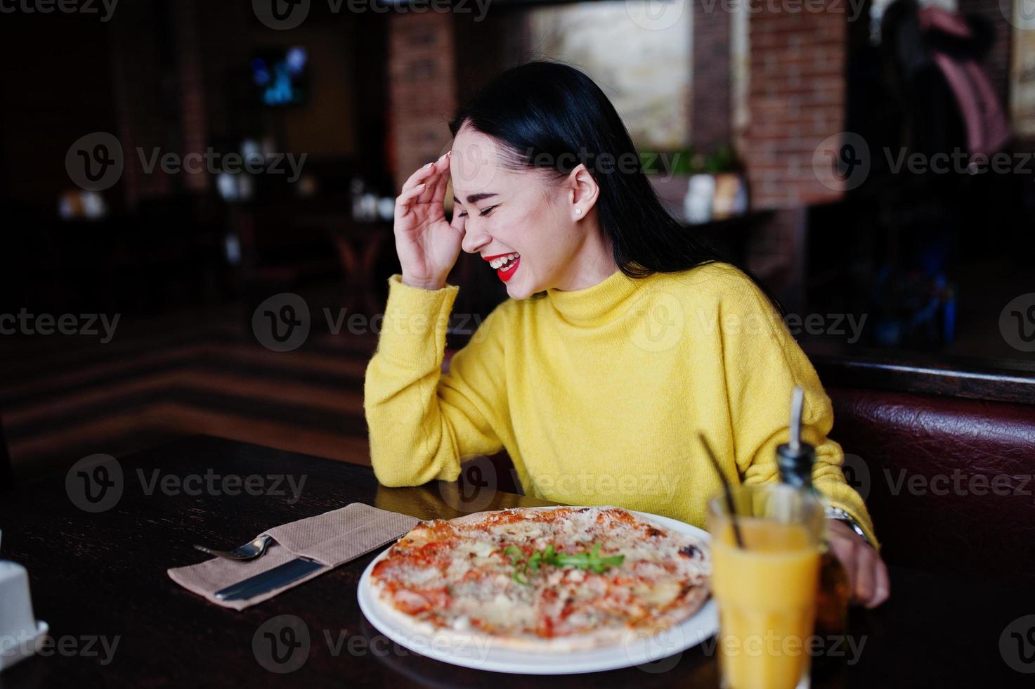 chica morena divertida en suéter amarillo comiendo pizza en el restaurante. foto