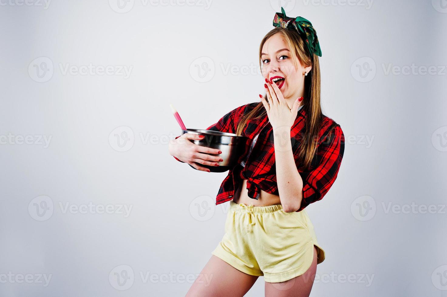 Young funny housewife in checkered shirt and yellow shorts pin up style with saucepan and kitchen spoon isolated on white background. photo