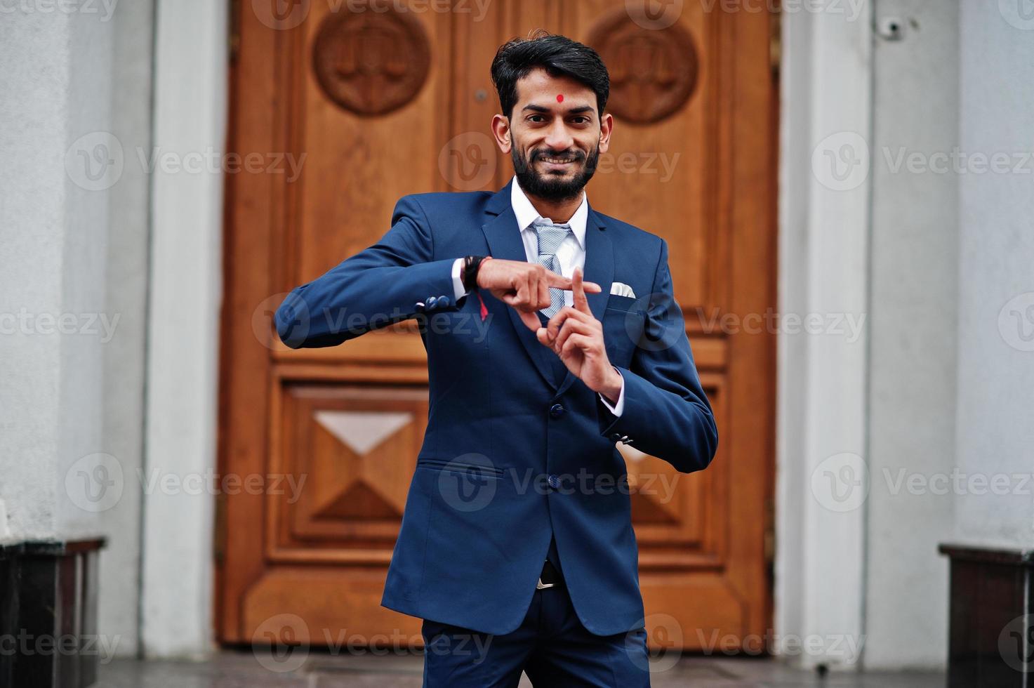 Stylish beard indian man with bindi on forehead, wear on blue suit posed outdoor against door of building and show cross at fingers. photo