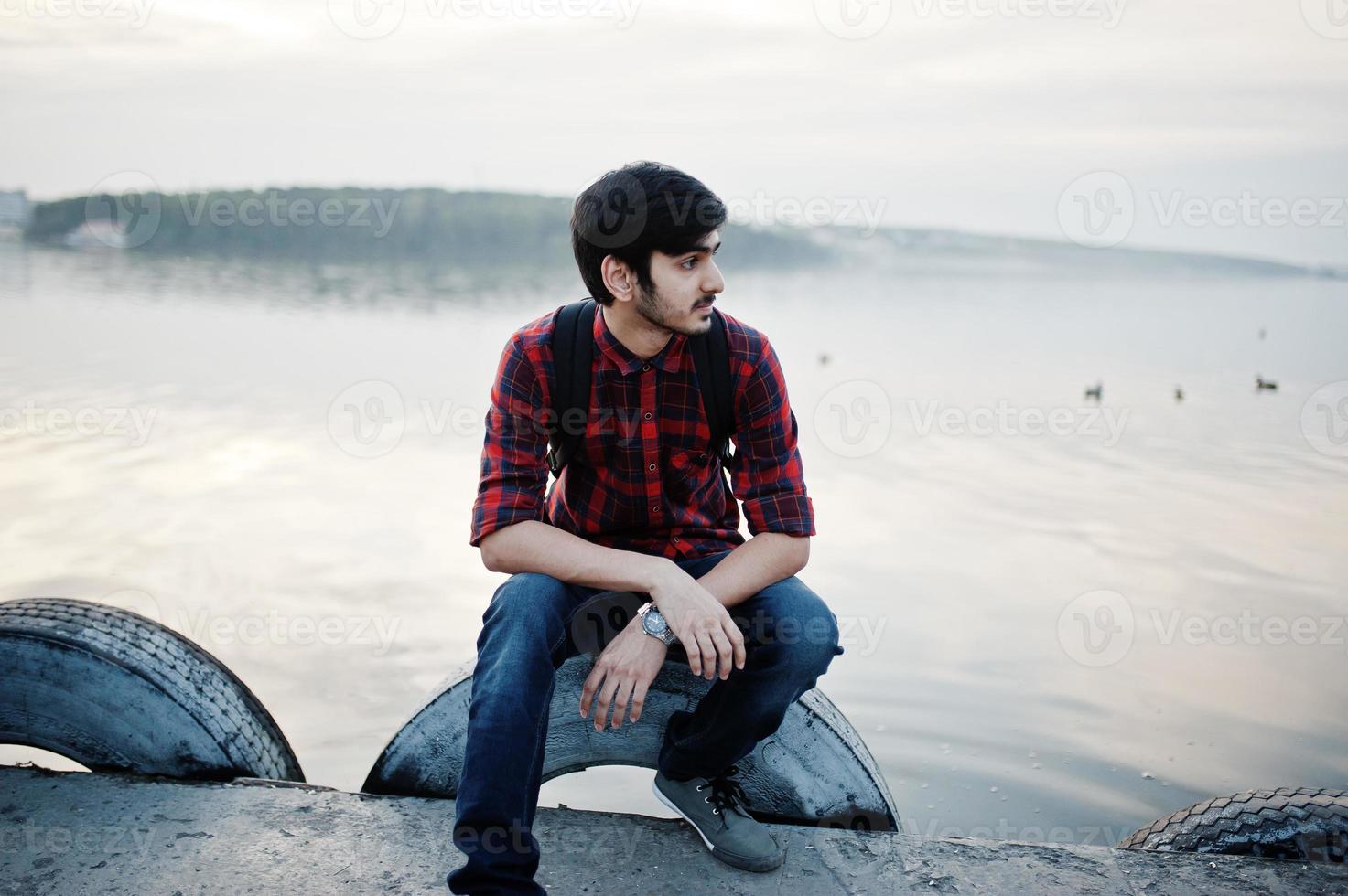 Young indian student man at checkered shirt and jeans with backpack posed on evening city against lake. photo