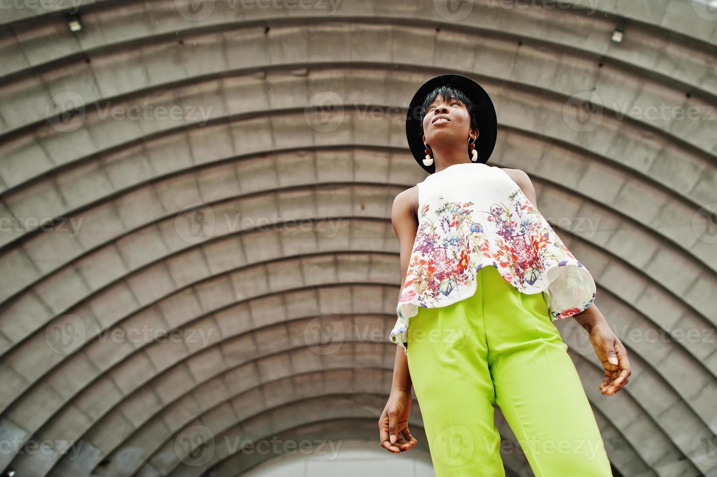 Amazing african american model woman in green pants and black hat posed outdoor against arena hall. photo