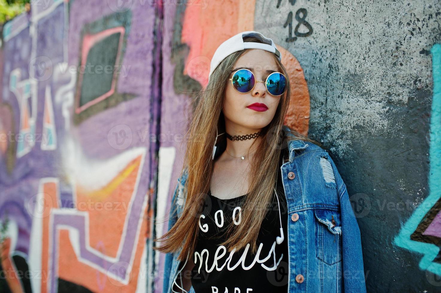 Stylish casual hipster girl in cap, sunglasses and jeans wear, listening music from headphones of mobile phone against large graffiti wall. photo