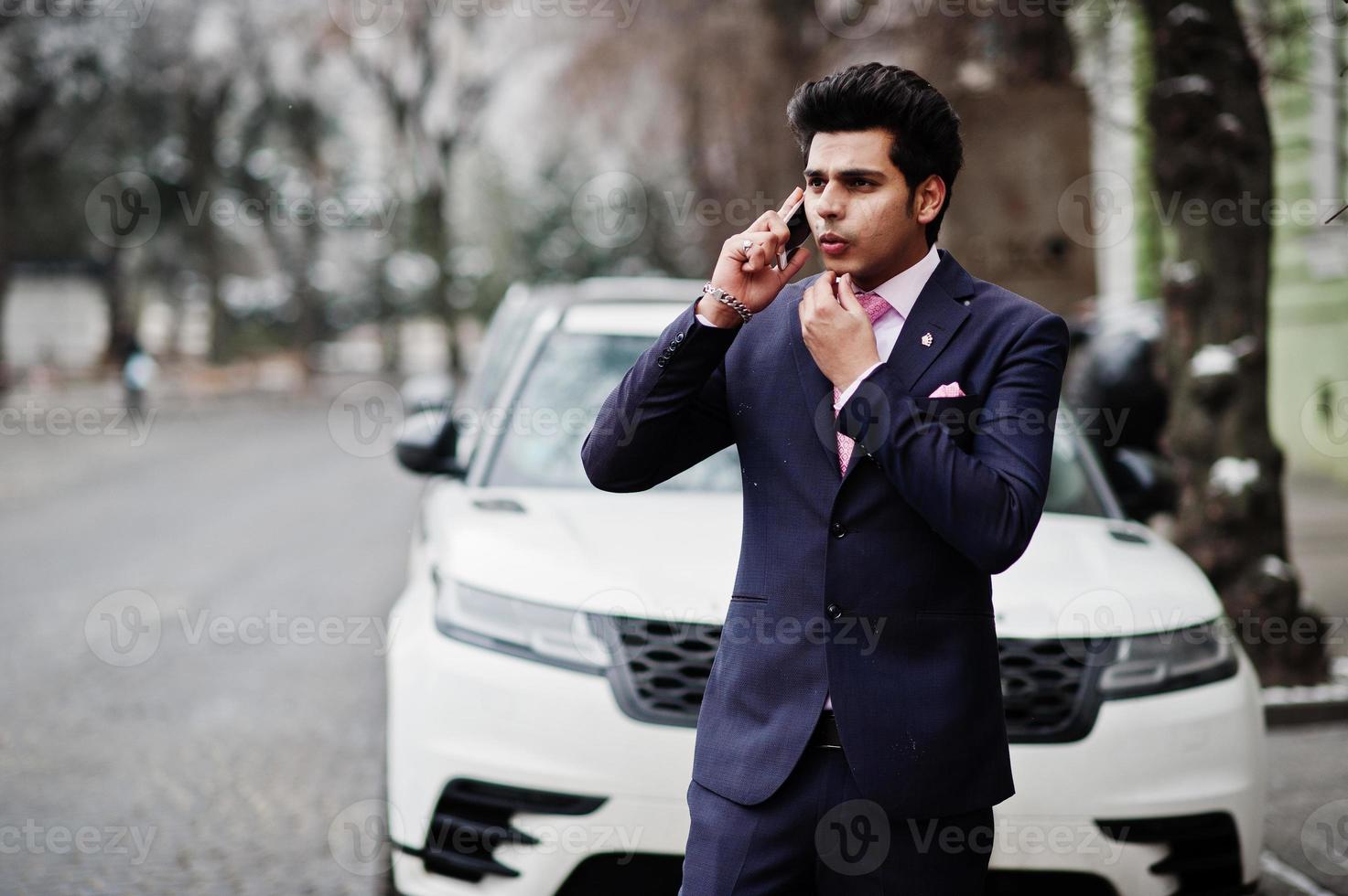 Elegant indian macho man model on suit and pink tie posed against white business car and speaking on phone. photo