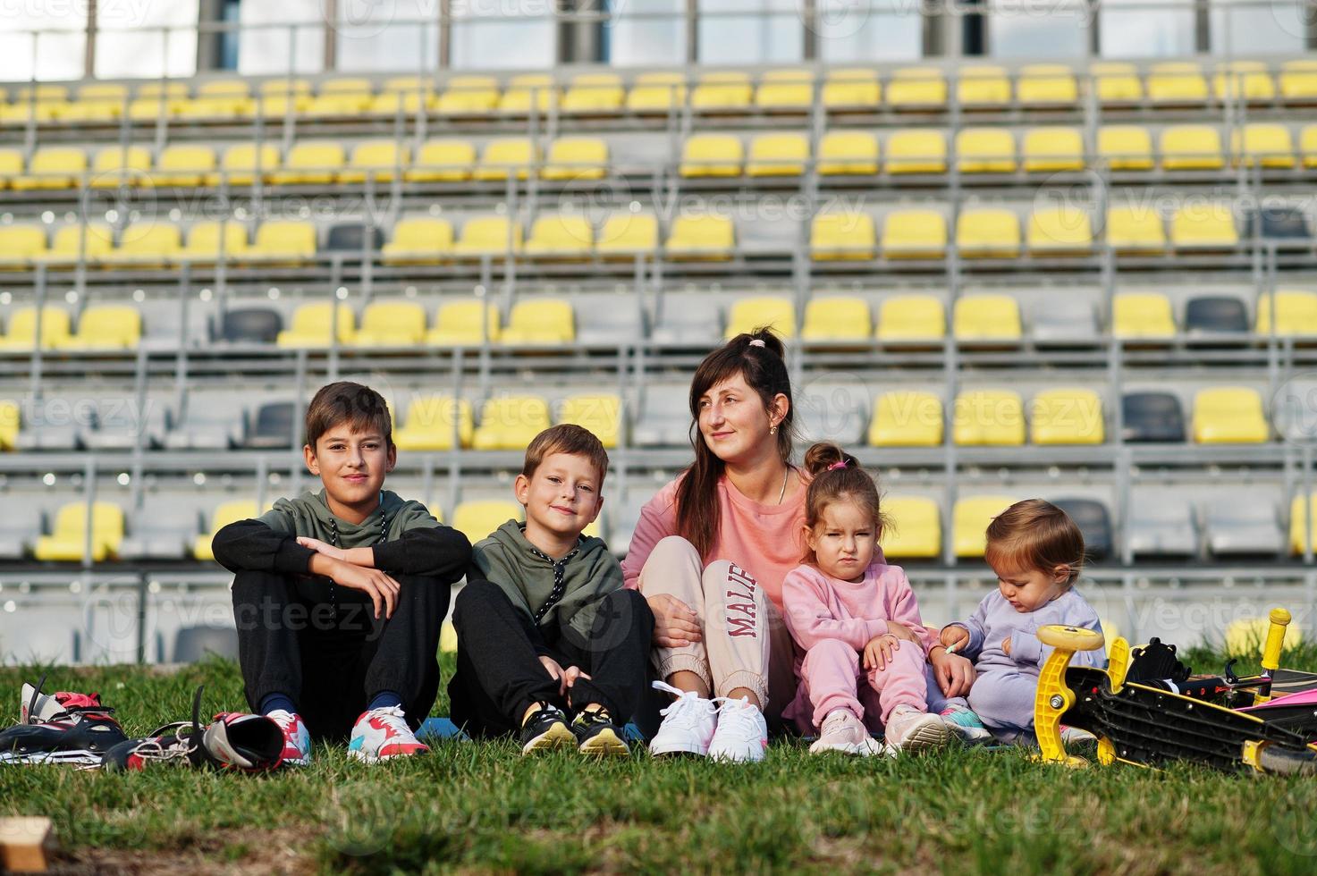 Young stylish mother with four kids sitting on grass against stadium. Sports family spend free time outdoors with scooters and skates. photo