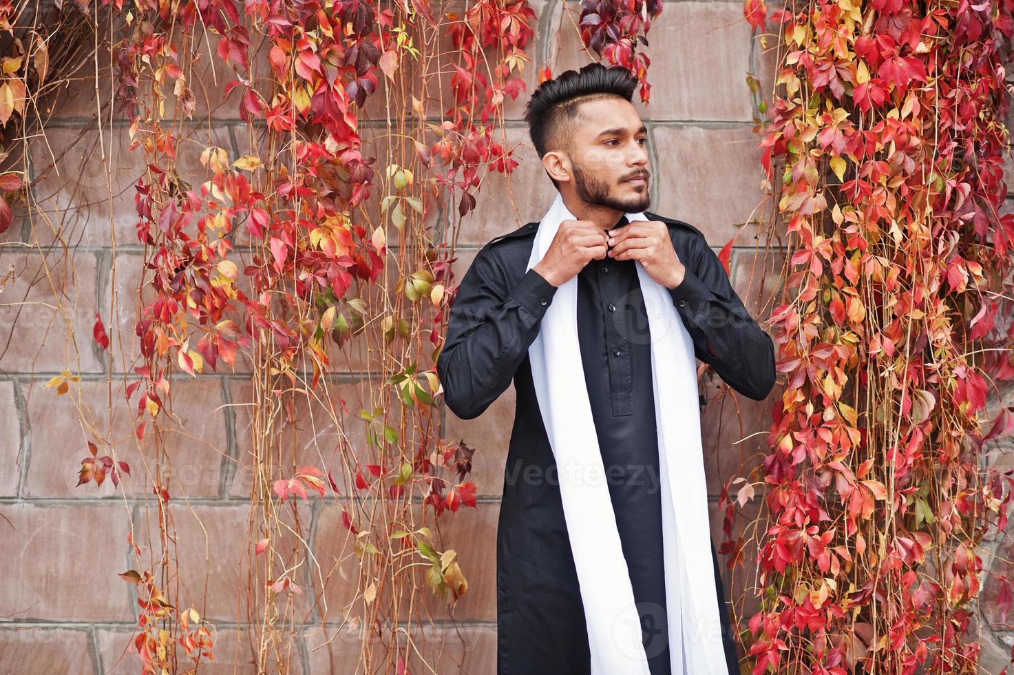 Indian stylish man in black traditional clothes with white scarf posed outdoor against red leaves wall. photo