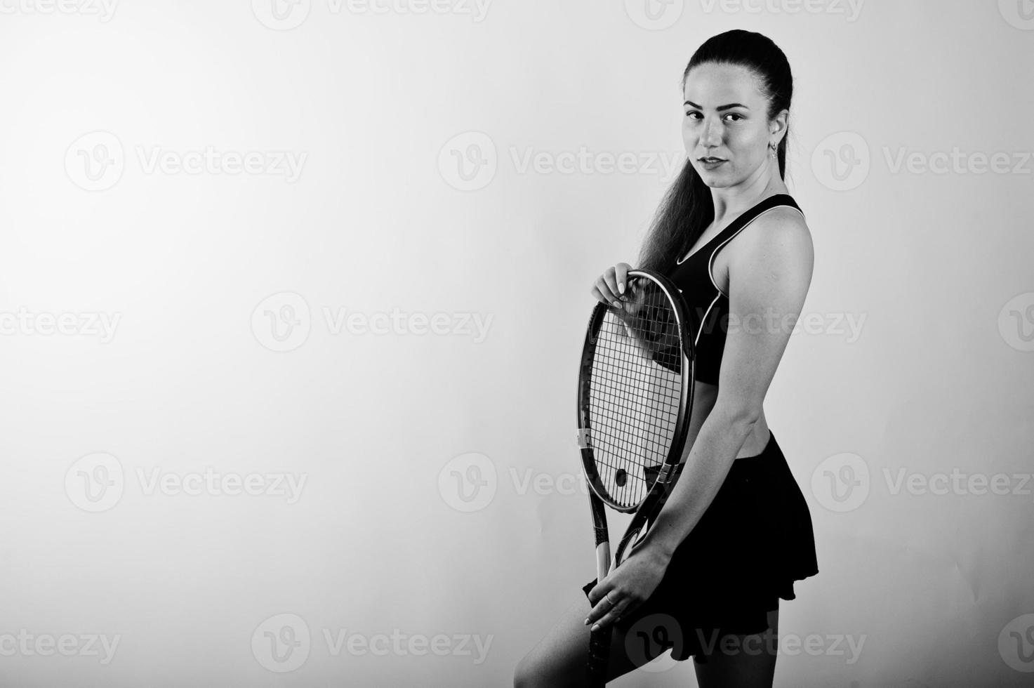 Black and white portrait of beautiful young woman player in sports clothes holding tennis racket while standing against white background. photo