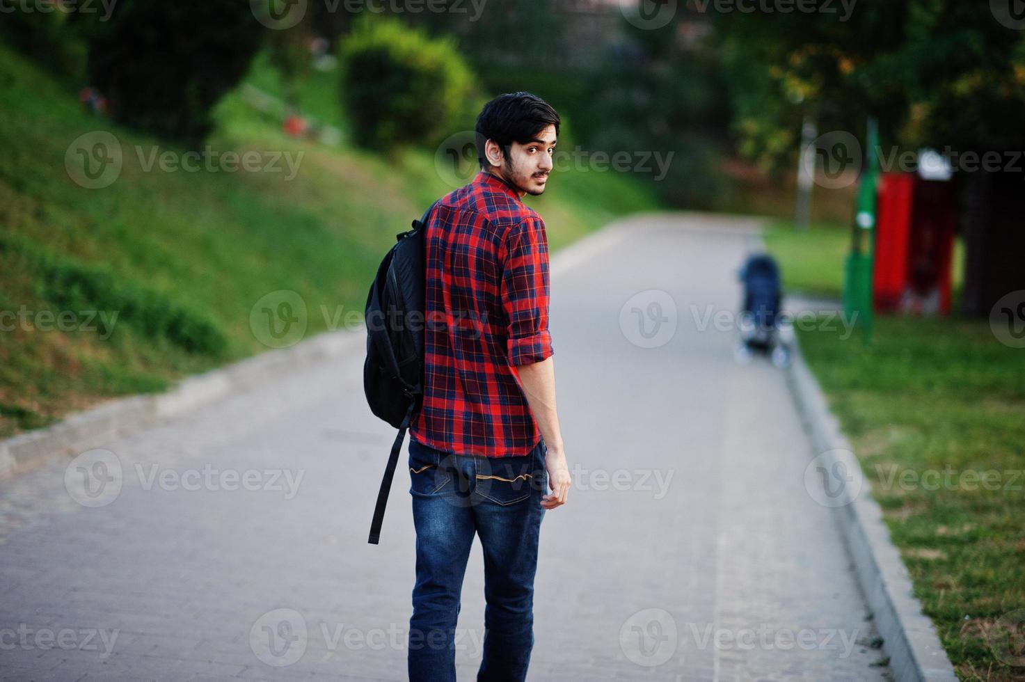 joven estudiante indio con camisa a cuadros roja y jeans con mochila posada en la calle. foto
