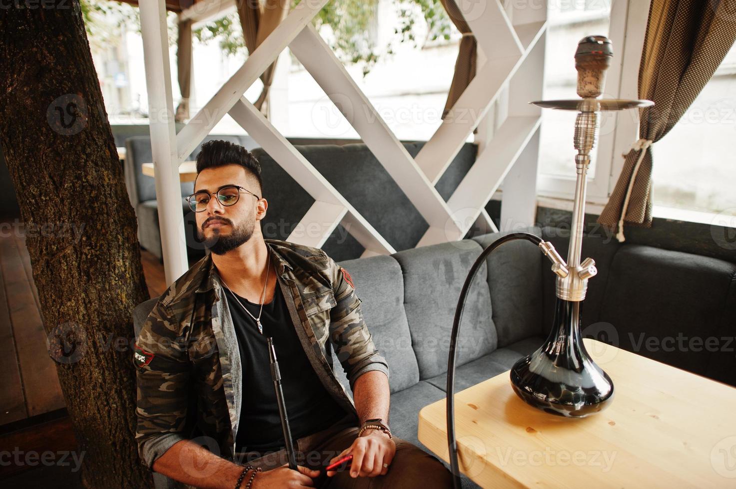 hombre árabe de barba elegante con gafas y chaqueta militar fumando narguile en el bar de la calle. modelo árabe descansando. foto