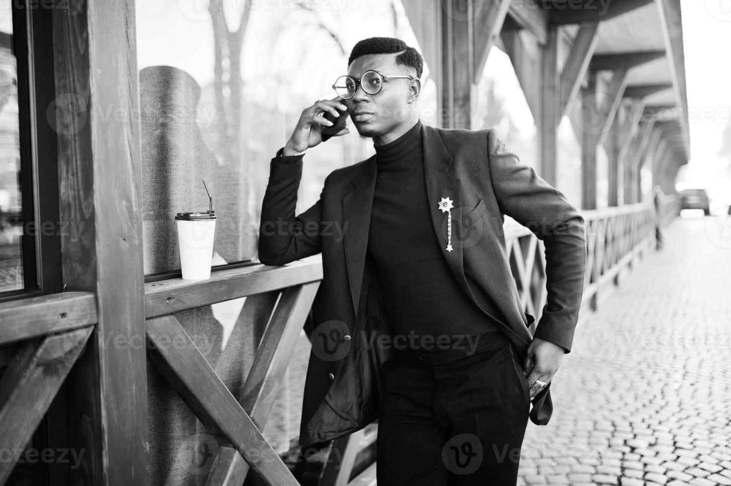 Amazingly looking african american man wear at blue blazer with brooch, black turtleneck and glasses posed at street. Fashionable black guy speaking on mobile phone. photo