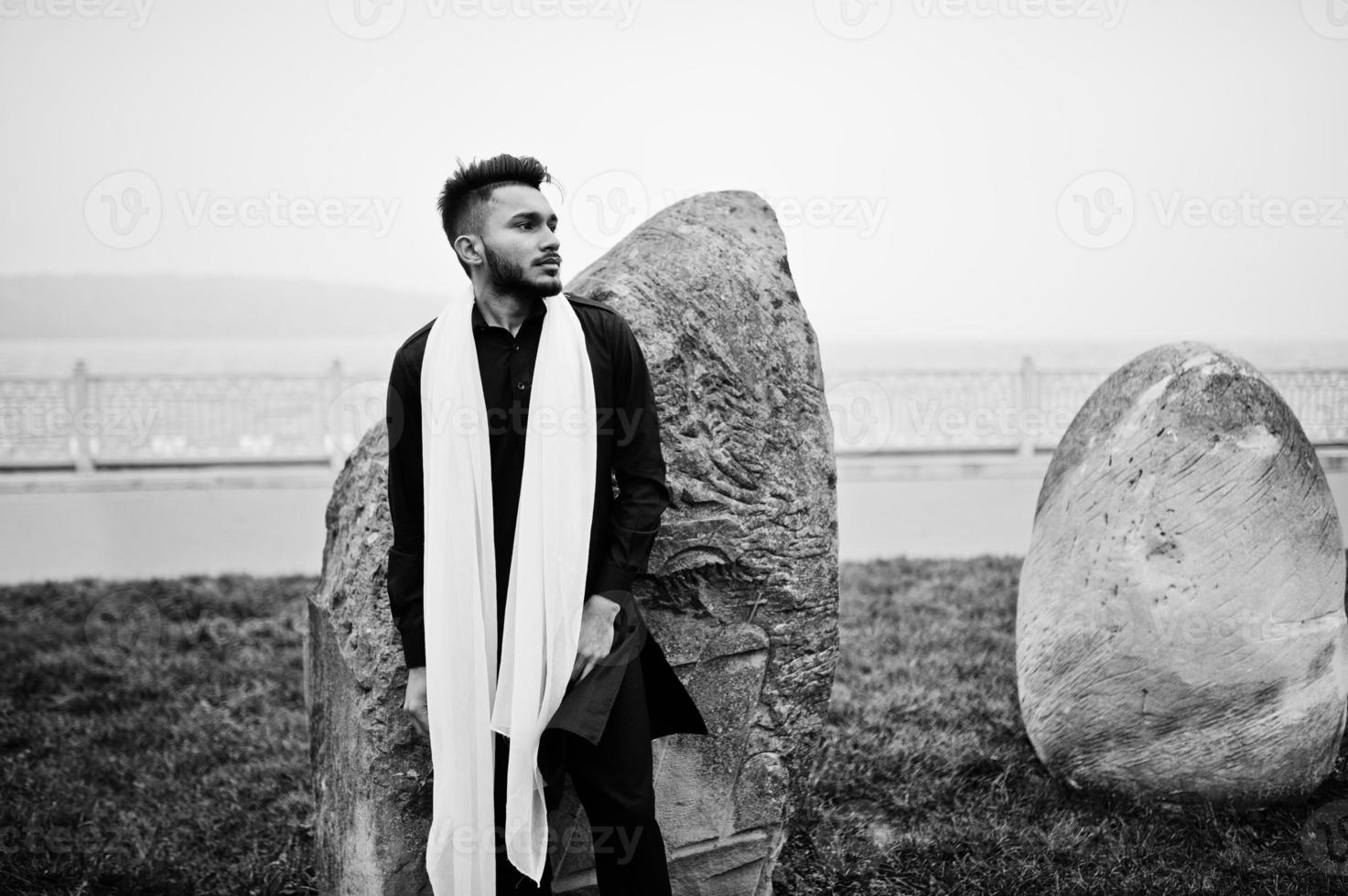 hombre indio con estilo en ropa tradicional negra con bufanda blanca posada al aire libre contra grandes piedras. foto