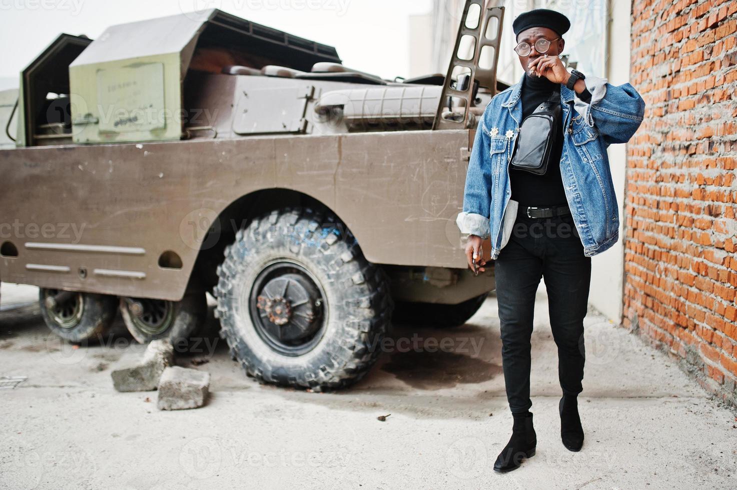 African american man in jeans jacket, beret and eyeglasses, smoking cigar and posed against btr military armored vehicle. photo