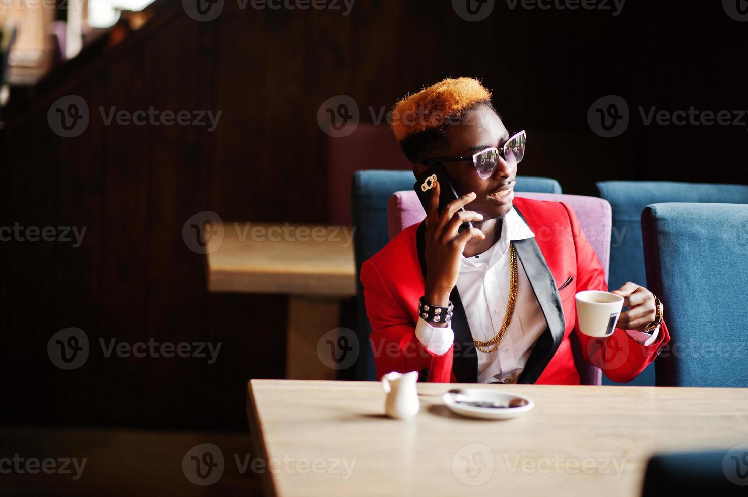 Fashion african american man model at red suit, with highlights hair and sunglasses sitting at cafe with coffee and speaking on mobile phone. photo