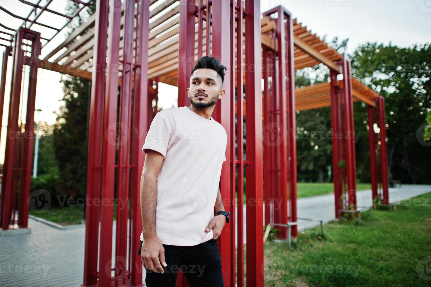 Stylish indian beard man at pink t-shirt. India model posed under red arch at park of city. photo