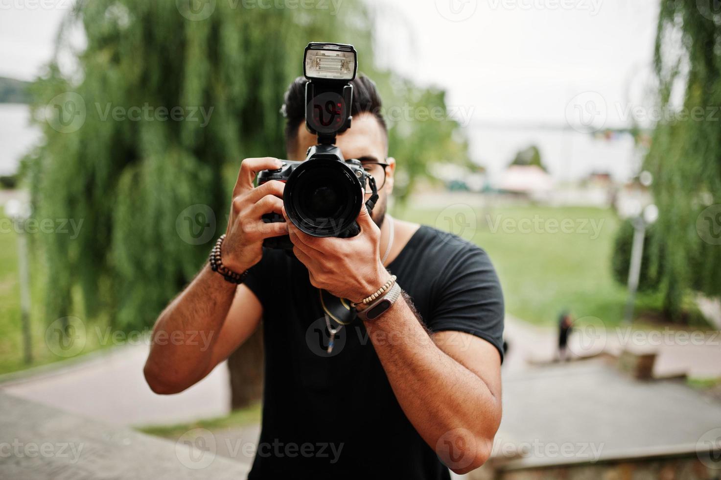 Impresionante y hermoso fotógrafo de hombre macho con barba árabe alta con gafas y camiseta negra con cámara profesional en las manos. foto