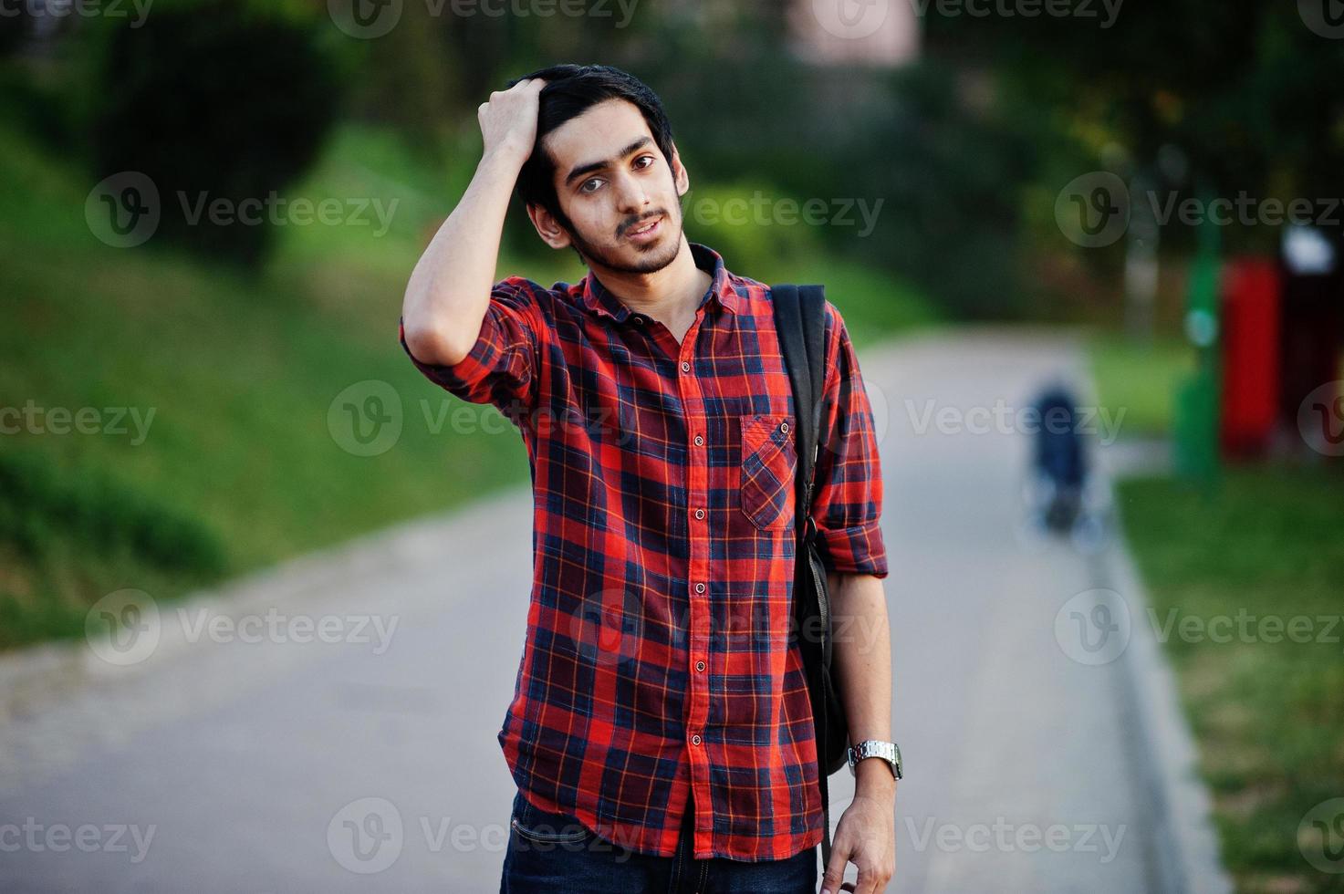 joven estudiante indio con camisa a cuadros roja y jeans con mochila posada en la calle. foto