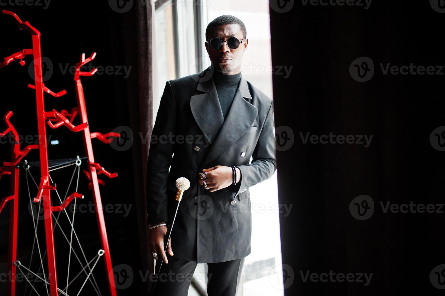 elegante caballero afroamericano con elegante chaqueta negra y gafas de sol, sosteniendo un bastón retro como matraz de caña o bastón basculante con mango de bola de diamante dorado. rico hombre afro de moda. foto