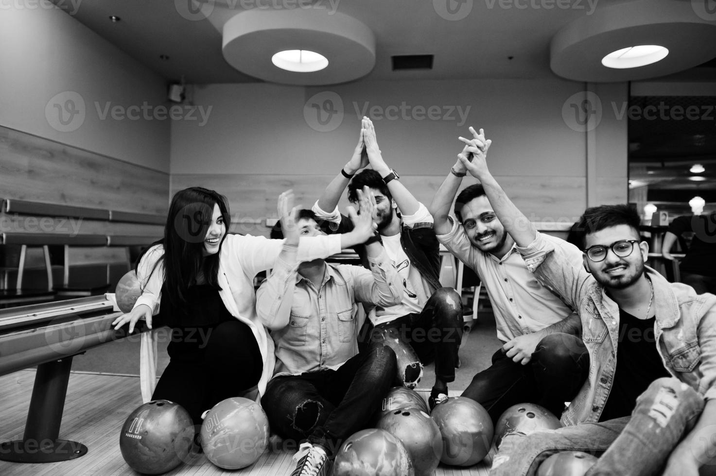 Group of five south asian peoples having rest and fun at bowling club. Holding bowling balls, sitting on alley and giving high five by hands. photo