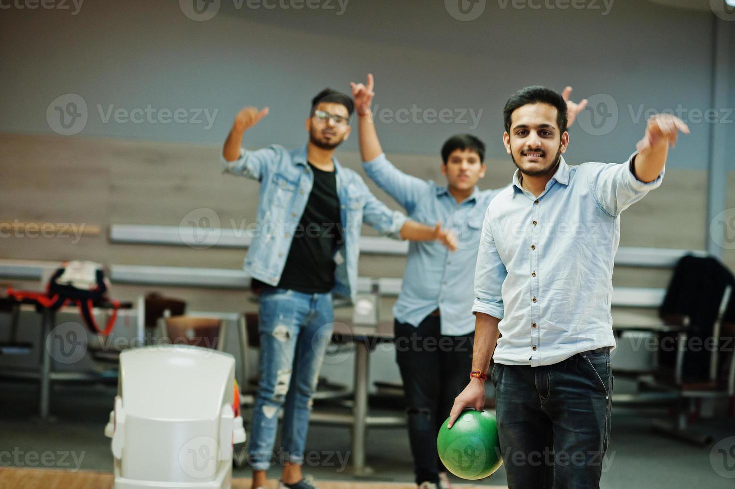 South asian man in jeans shirt standing at bowling alley with ball on hands. Guy is preparing for a throw. Friends support him loudly. photo