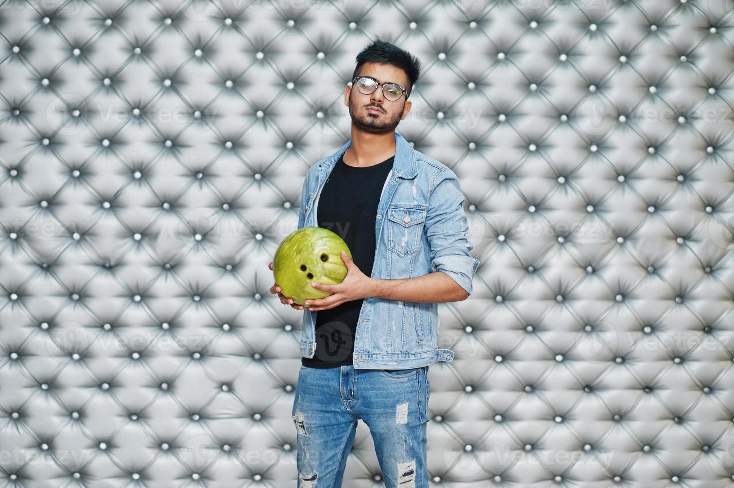 Stylish asian man in jeans jacket and glasses with bowling ball at hand against silver wall background. photo