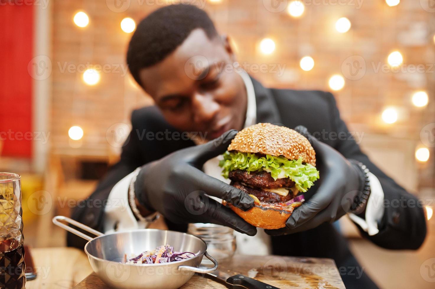 Respectable young african american man in black suit and gloves for food sitting in restaurant with tasty double burger and soda drink. photo