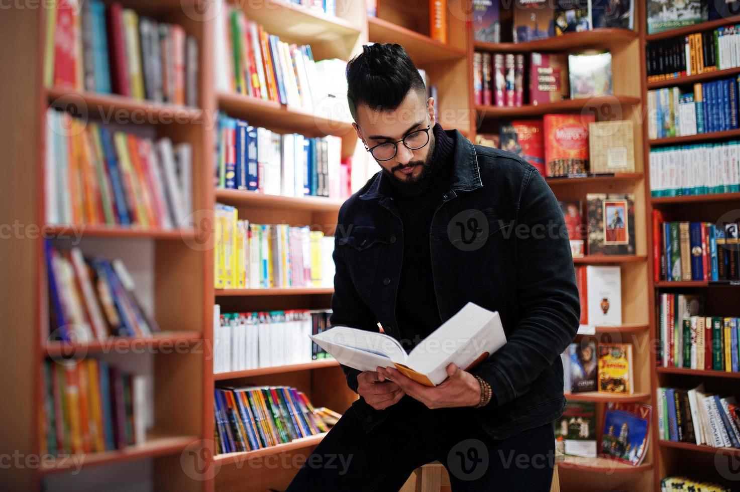 Tall smart arab student man, wear on black jeans jacket and eyeglasses, at library with book at hands. photo