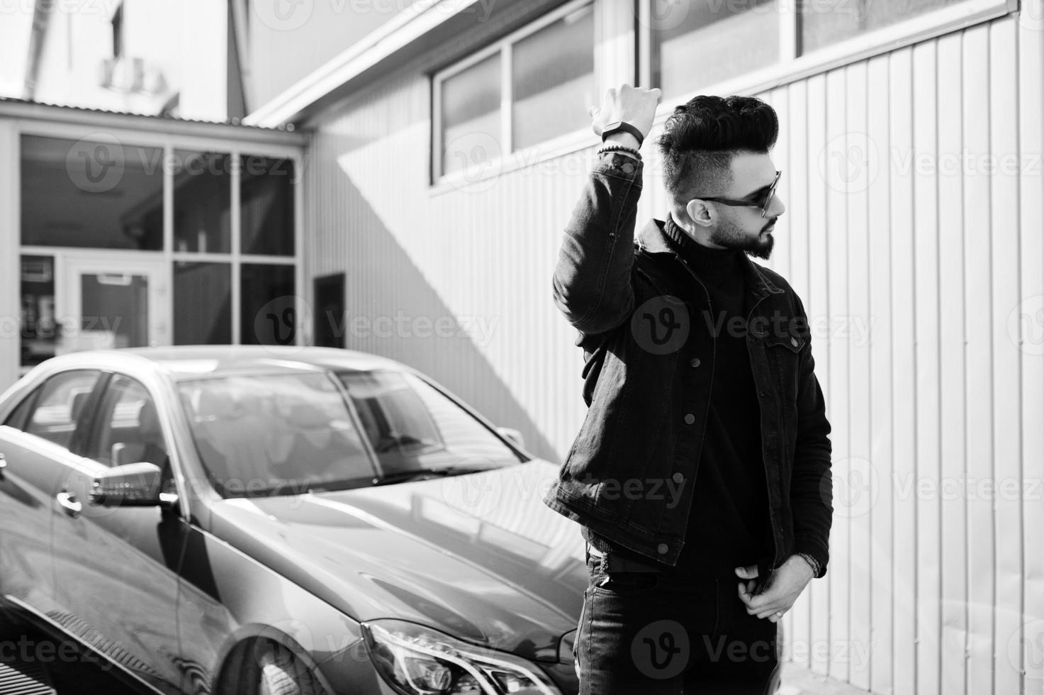 Fashion Arab man wear on black jeans jacket and sunglasses posed against business modern car. Stylish, succesful and fashionable arabian model guy. photo