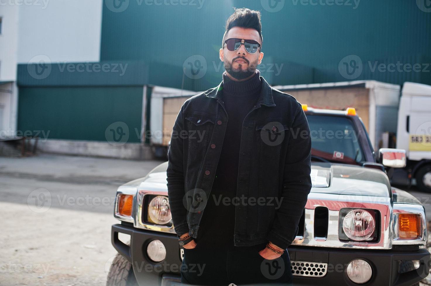 El hombre árabe de barba rica en moda usa una chaqueta negra de jeans y gafas de sol posadas contra un gran auto todoterreno negro. chico modelo árabe elegante, exitoso y de moda. foto